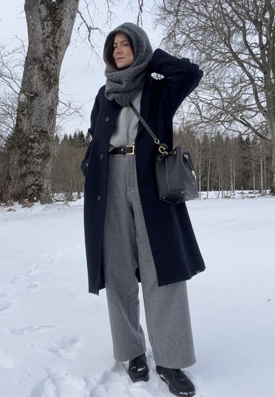 Mujer con abrigo de invierno, bufanda, pantalones anchos grises y botas negras, de pie sobre la nieve cerca de árboles deshojados, con un bolso negro colgado del hombro.