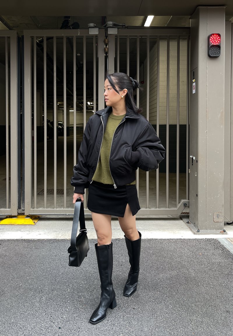 Veste bomber noire portée sur un pull vert olive, assortie à une mini-jupe noire avec une fente latérale et des bottes noires à talons carrés montant jusqu'aux genoux.