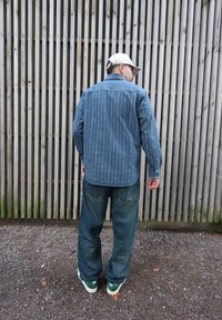 Chemise en denim bleu à rayures verticales, associée à un jean bleu foncé ample et des baskets vertes. L'arrière-plan est composé de lattes en bois.