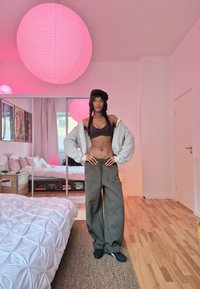 Sweat à capuche gris surdimensionné, top court marron et pantalon large olive. La pièce comprend un sol en bois, une housse de lit blanche et des lanternes en papier roses.