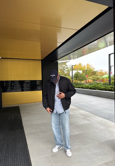 Chaqueta bomber negra sobre una camisa a rayas azules y blancas, jeans azul claro y zapatillas blancas con rayas negras; el fondo presenta paneles amarillos y negros.