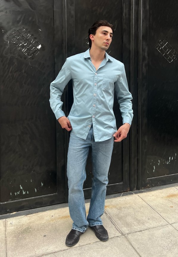 Ung mand står ved en sort metal dør iført en lyseblå skorte med knapper, blå jeans og sorte loafers.