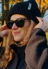 Juodas mezgintas kepurės bžūp su etikete, tamsūs akiniai nuo saulės ir juoda tekstūruota striukė. Dalinai matomi minkšti, banguoti plaukai. Saulėta fonas su geltonais lapais.