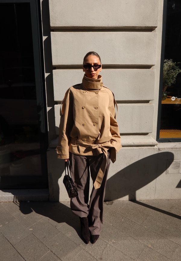 Vrouw met een beige trenchcoat met hoge kraag, bruine broek, zonnebril en een zwarte handtas vasthoudend, staand tegen een stenen muur.