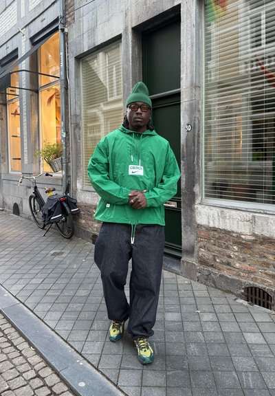 Chaqueta verde con capucha y cordones, pantalones anchos oscuros y zapatillas verde/amarillo; de pie en una calle de adoquines cerca de una bicicleta.