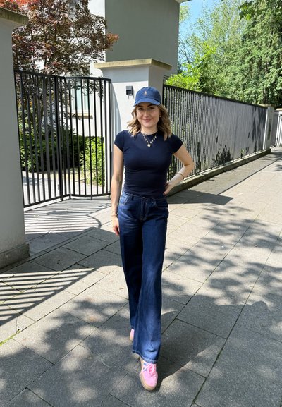 Camiseta ajustada de la marina, jeans de pierna ancha oscuros y zapatillas rosas. Gorra con logo. Entorno al aire libre con vegetación y sombras de luz solar.