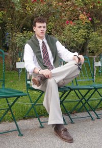 Męska postać siedzi na zielonym krześle, ubrana w białą koszulę, kratkowany krawat, oliwkowy sweter zarzucony na ramiona, beżowe spodnie i brązowe buty.