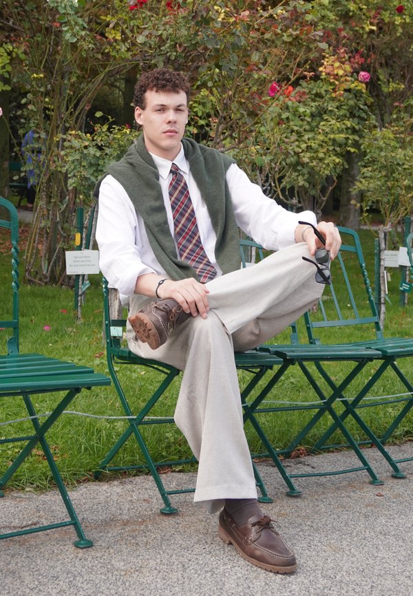 Eine männliche Person sitzt auf einem grünen Stuhl, trägt ein weißes Hemd, eine karierte Krawatte, einen olivfarbenen Pullover über den Schultern, beigefarbene Hose und braune Schuhe.
