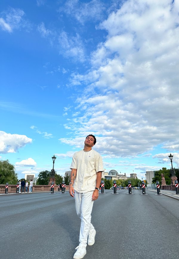 Weißes oversize Hemd und weiße Hose, die auf einer Straße gehen. Blauer Himmel mit Wolken im Hintergrund. Barrieren säumen die Straße, Stadtsilhouette sichtbar.