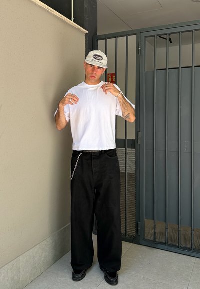 Hombre vestido con una camiseta blanca, pantalones negros con cadena, zapatos negros y una gorra blanca, de pie cerca de una puerta gris con barrotes en el interior.