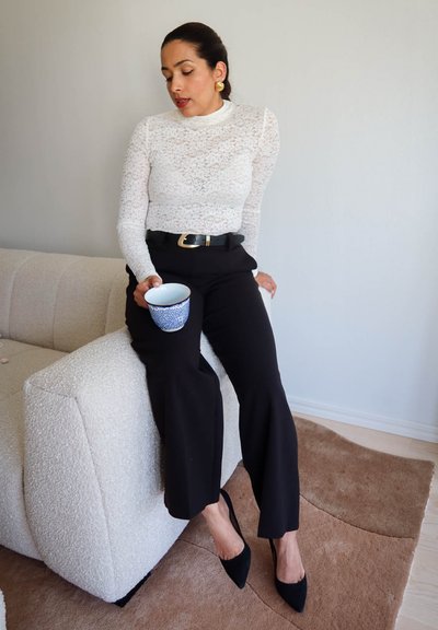 Mujer con blusa blanca de encaje y pantalones negros sentada en el reposabrazos del sofá, sosteniendo una taza con estampado azul, mirando hacia abajo con los ojos cerrados.