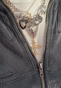 Collares en capas con corazones de plata, una cruz con acentos de color y una cadena texturizada, usados sobre una camisa blanca y una chaqueta negra con cremallera.