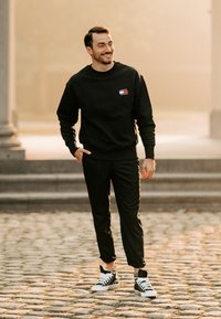 Fekete kapucnis pulóver, amelyen egy kis színes logó található a mellrészen, fekete nadrággal és fekete-fehér sportcipőkkel párosítva, macskaköves járdán.