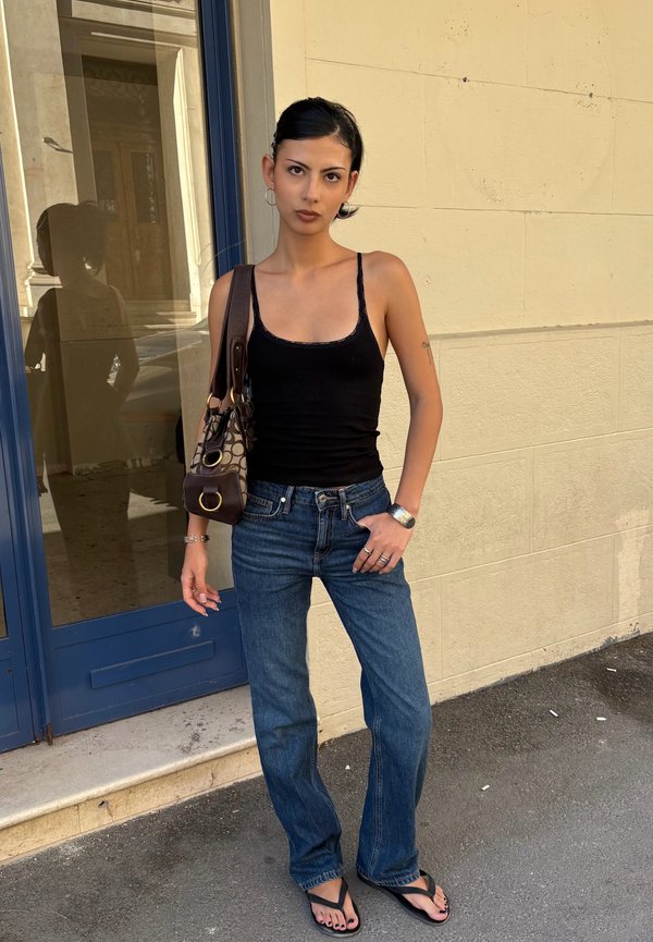 Mujer joven con camiseta sin mangas negra, vaqueros azules y sandalias, de pie junto a una pared beige y una puerta de vidrio, llevando un bolso con estampado en el hombro.