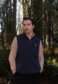 Veste sans manches marine avec manches en polaire beige, dotée d'une fermeture éclair et d'un petit logo sur la poitrine. Présentée sur un fond de forêt verdoyante.