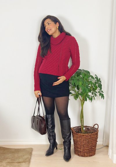Mujer con cabello largo y oscuro que lleva un suéter de punto rojo, falda negra, medias negras y botas hasta la rodilla, sosteniendo un bolso marrón junto a una planta en maceta verde.