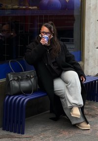 Sort frakke over en sort hættetrøje, grå joggingbukser og beige sko. Holder en blå kaffekop, siddende på en blå bænk ved siden af en sort håndtaske.