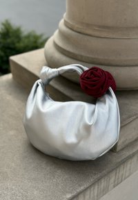 Mjuk silverhandväska med ett knutet handtag och en röd tygblomma, som vilar på en stenplatta nära en kolonn utomhus.