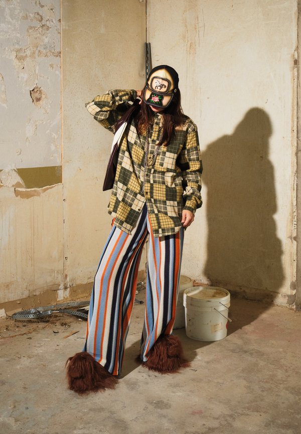 Persona que lleva una chaqueta de cuadros amarilla, pantalones a rayas, zapatillas peludas y una gorra decorada, de pie en una habitación desgastada y sin terminar con cubos de pintura.