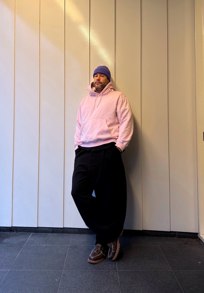 Homme en sweat à capuche rose, bonnet bleu, pantalon large noir et bottes marron, appuyé contre un mur crème à panneaux à l'intérieur.