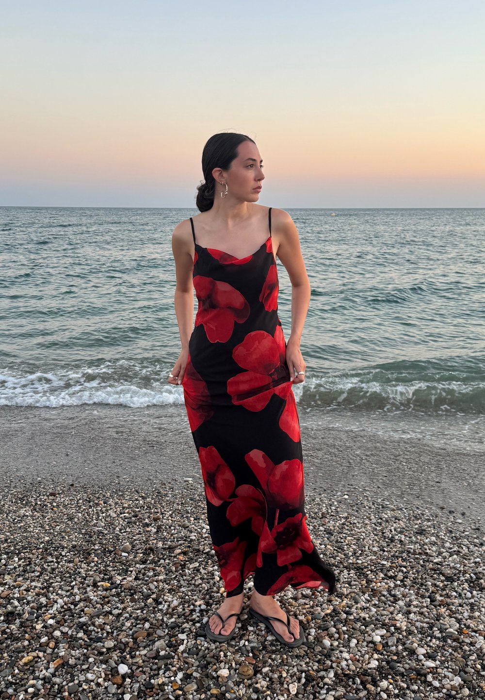 Zwarte jurk met rood bloemenpatroon, dunne bandjes, getailleerd ontwerp, gedragen met zwarte teenslippers, staande op een kiezelstrand bij de zee.