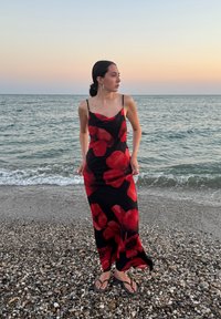 Zwarte jurk met rood bloemenpatroon, dunne bandjes, getailleerd ontwerp, gedragen met zwarte teenslippers, staande op een kiezelstrand bij de zee.