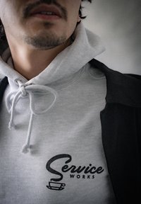 Szara bluza z kapturem i sznurkiem w okolicy szyi, z czarnym logo "Service Works" przedstawiającym ikonę filiżanki kawy, umieszczona na tle ciemniejszej kurtki.