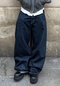 Tamne traper hlače širokih nogavica s vidljivim detaljima šavova, uparene s crnim čizmama s debelim đonom. Tkanina izgleda teška i teksturirana.