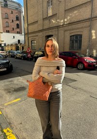 Szőtt narancssárga kézitáska két pánttal, szürke, fél vállig érő pulóver és szürke farmernadrág. Városi környezet, téglaépületekkel és parkoló autókkal.