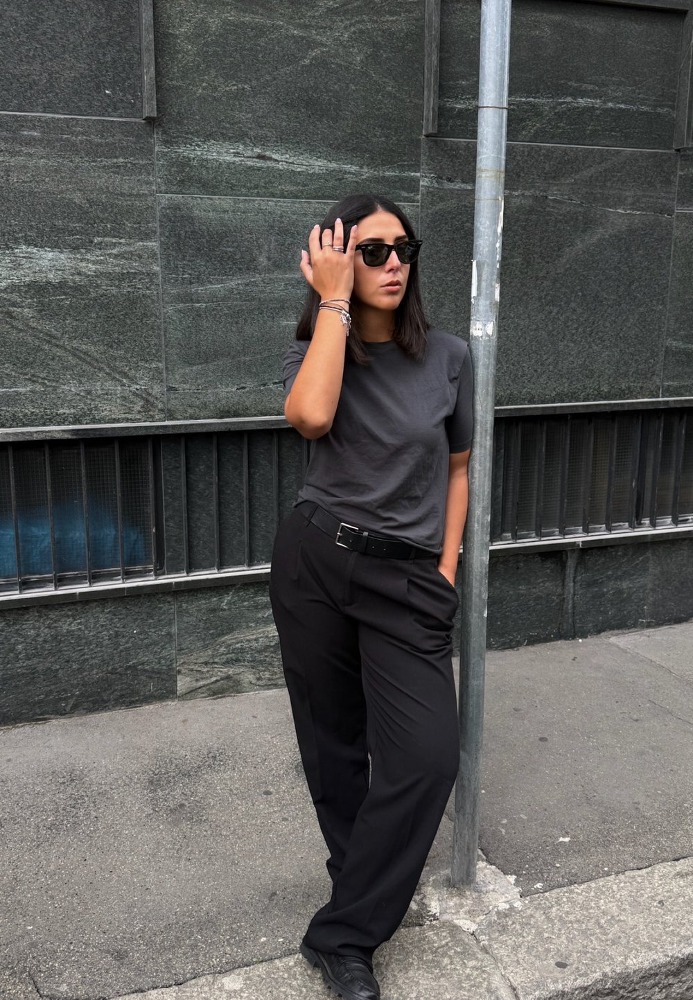 Kvinne med skulderlangt mørkt hår som har på seg solbriller, grå t-skjorte og svarte bukser, står og lener seg mot en metallstang på fortauet.