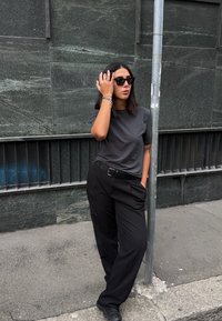 Kvinne med skulderlangt mørkt hår som har på seg solbriller, grå t-skjorte og svarte bukser, står og lener seg mot en metallstang på fortauet.