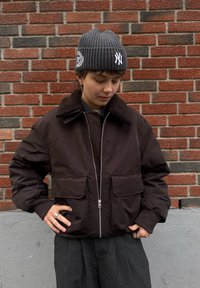 Giacca bomber marrone con colletto in shearling, chiusura con zip e due tasche frontali. Indossata con un berretto grigio a coste. Sfondo in mattoni rossi.