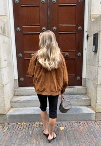 Brun semsket oversized jakke, svarte capri leggings og flip-flops. Bærer en brun mønstrete håndveske. Står på steintrapper.