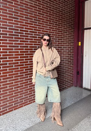 Mujer con chaqueta beige extragrande, pantalones cortos de mezclilla claros, botas marrones con flecos, gafas de sol y sosteniendo un bolso con estampado frente a una pared de ladrillos.