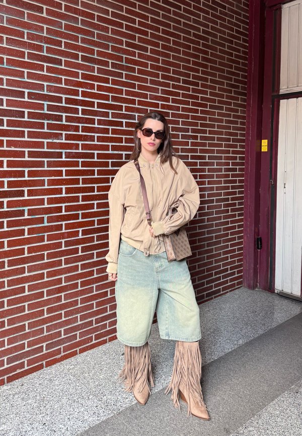 Donna che indossa una giacca beige oversize, shorts in denim chiaro, stivali color cuoio con frange, occhiali da sole e tiene una borsa con motivo davanti a un muro di mattoni.