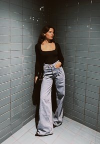 Femme aux longs cheveux bruns portant un haut noir à manches longues et un jean bleu à jambes larges, debout contre des murs carrelés bleus sous un éclairage doux.