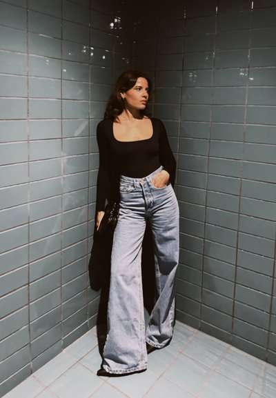 Mujer con cabello largo y oscuro, vestida con una blusa negra de manga larga y vaqueros azules de pierna ancha, de pie contra paredes azulejadas en azul bajo una iluminación suave.