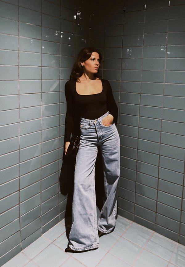 Femeie cu păr lung și închis la culoare, purtând un top negru cu mâneci lungi și blugi largi de culoare albastră, stă în fața unui perete cu plăci albastre, iluminată de o lumină blândă.