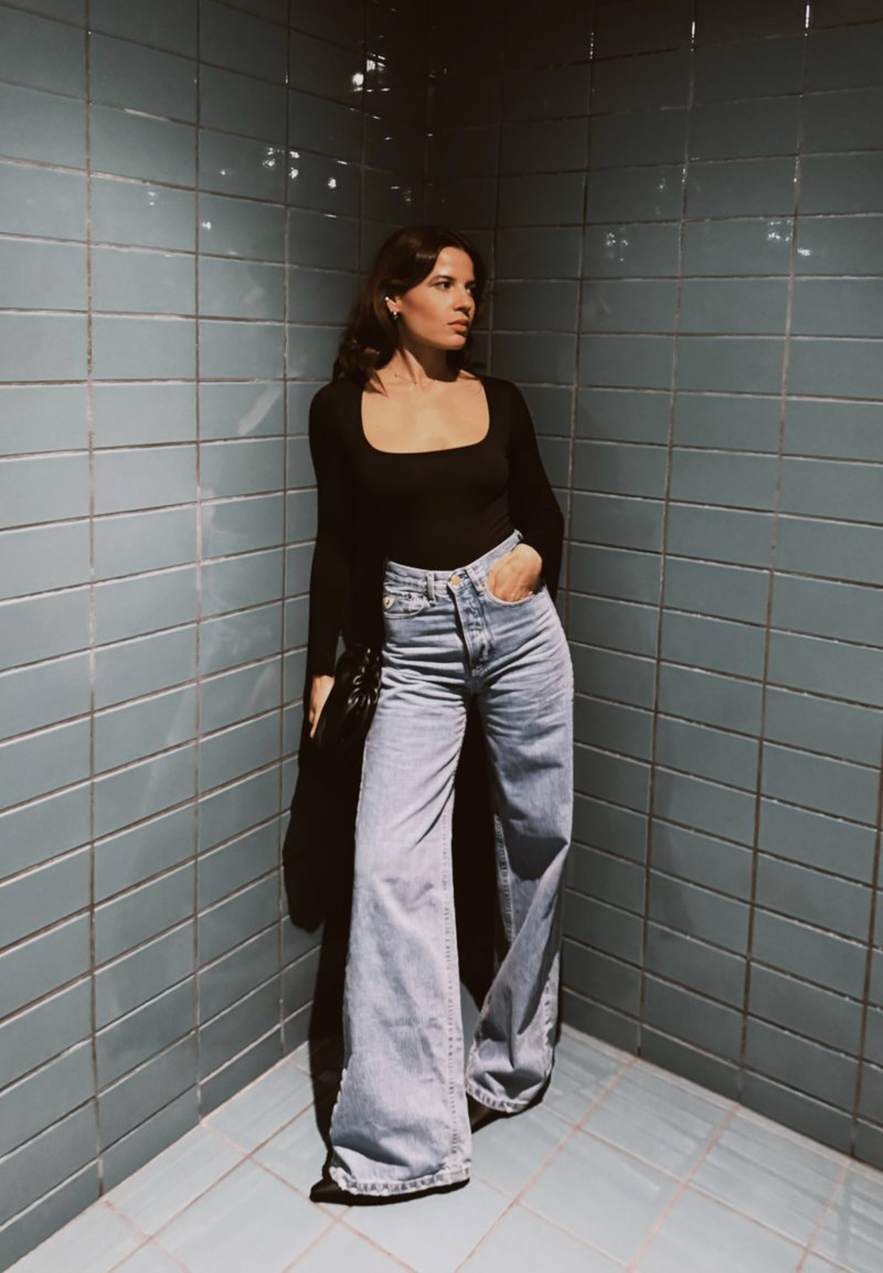 Femme aux longs cheveux bruns portant un haut noir à manches longues et un jean bleu à jambes larges, debout contre des murs carrelés bleus sous un éclairage doux.