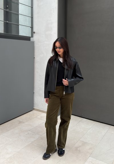 Mujer con chaqueta de cuero negra, top negro, pantalones oliva, zapatillas oscuras y gafas de sol, de pie en el interior junto a paredes grises y blancas.
