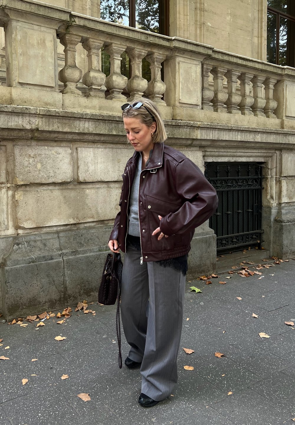 Femme portant une veste en cuir bordeaux surdimensionnée, un pantalon gris à jambes larges, un haut gris, tenant un sac à main foncé, marchant sur un trottoir de la ville jonché de feuilles.