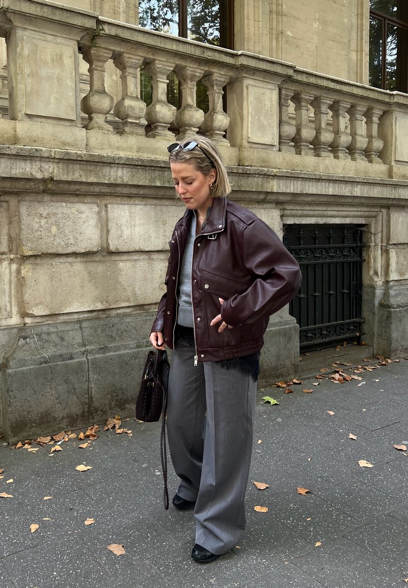 Femme portant une veste en cuir bordeaux surdimensionnée, un pantalon ample gris, un haut gris, tenant un sac à main foncé, marchant sur un trottoir de la ville jonché de feuilles.