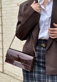 Personne portant un blazer marron sur une chemise blanche avec une jupe à carreaux, tenant un sac à main brun foncé brillant à bandoulière chaîne contre un mur en briques.