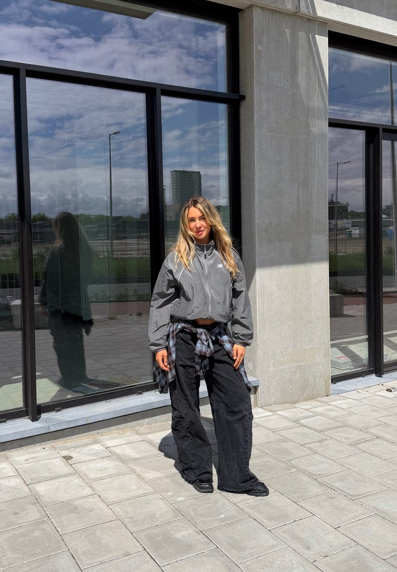 Blond kvinne står utenfor en moderne bygning med store vinduer, iført grå jakke, svarte vide bukser og rutete skjorte knyttet i livet.