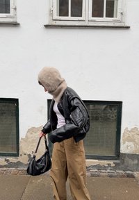 Personne portant une capuche beige duveteuse, une veste en cuir noire, une chemise blanche et un pantalon marron, marchant sur un trottoir en tenant un sac à main noir près d'un mur blanc.
