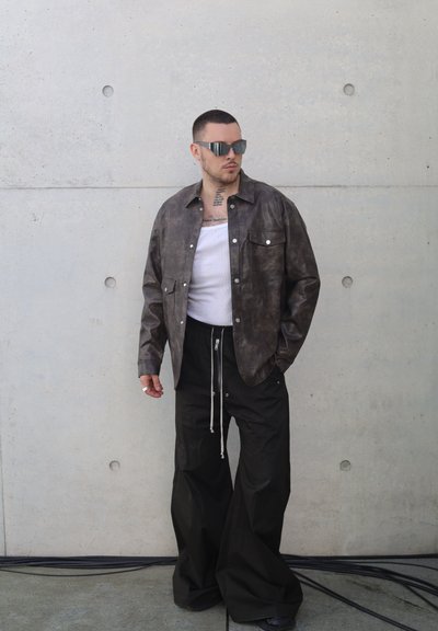 Chaqueta de cuero marrón con dos bolsillos frontales, llevada sobre una camiseta sin mangas blanca. Pantalones amplios negros con cordón. Gafas de sol delgadas y angulares.