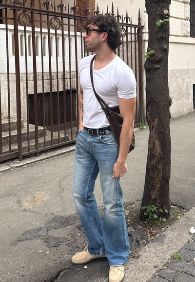 Camiseta blanca ajustada, vaqueros azules desgastados, zapatos beige tipo slip-on y un bolso marrón de hombro. El fondo presenta una valla de metal y un árbol.