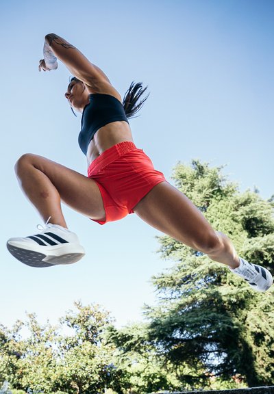Atleta con un top corto negro y pantalones cortos rojos salta contra un cielo despejado. Lleva zapatillas blancas con rayas negras. Tatuajes visibles en su brazo.