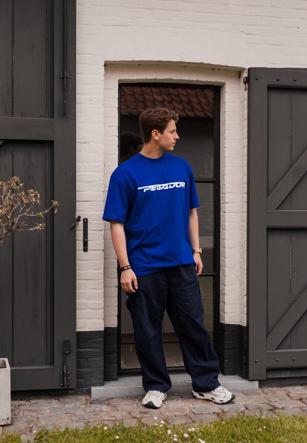 Camiseta de algodón azul con el logo "PEGAJOR" en blanco, de corte relajado. Combinada con pantalones sueltos azul marino y zapatillas blancas. De pie en el marco de la puerta.