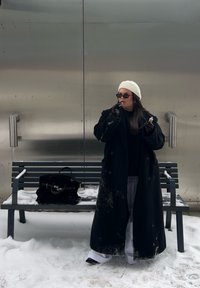 Femme en manteau noir, béret blanc et gants appliquant du rouge à lèvres en se tenant dans la neige près d'un banc avec un sac noir contre un mur en métal.